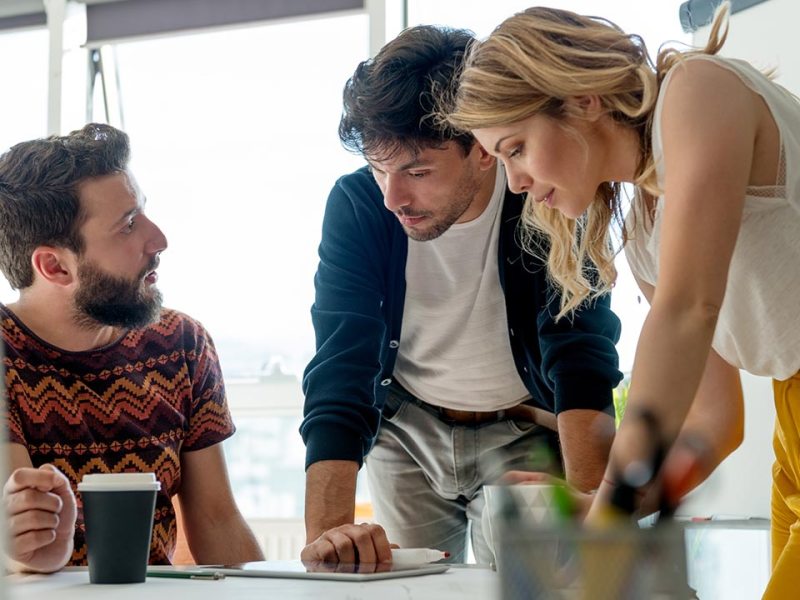 Three coworkers lean over a desk talking about work