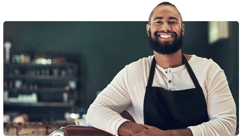 Barber in an apron smiles at the camera and leans on his chair.