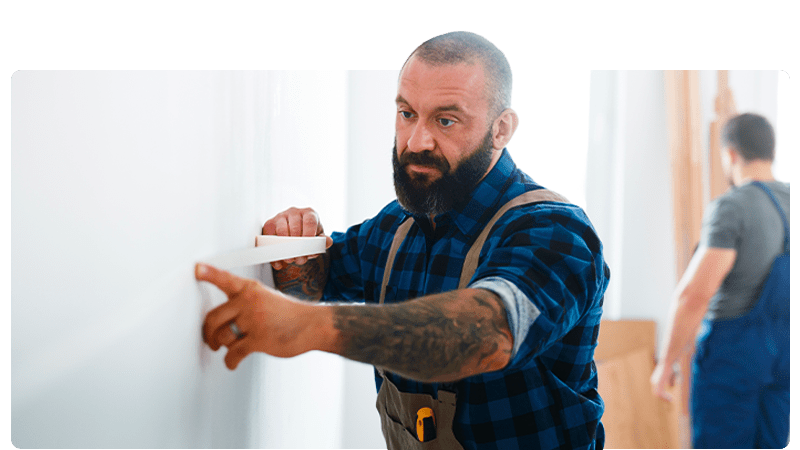 Bedroom fitter in overalls and a blue shirt masking a wall while a man works behind him.